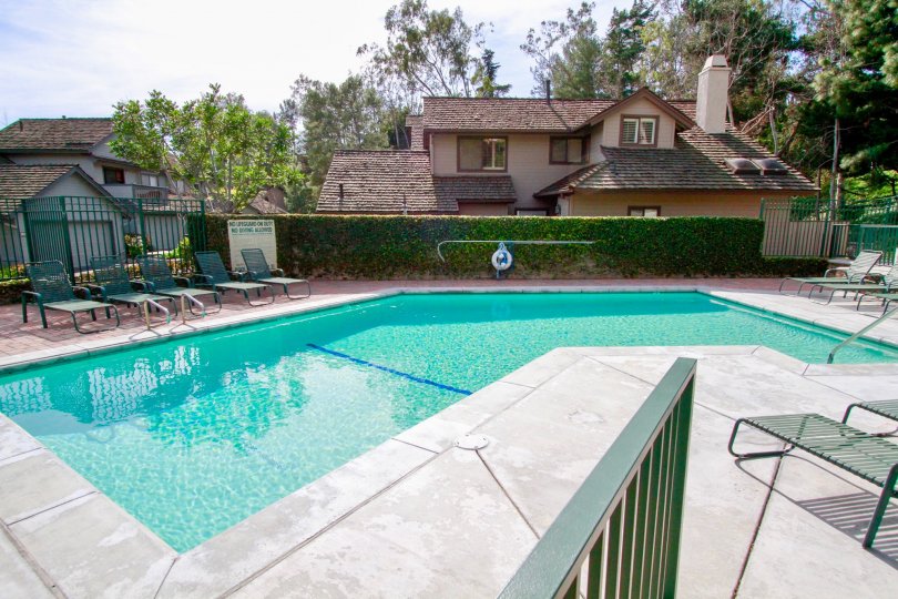 THE SWIMMING POOL WITH SITTING CHAIRS AND THE ENTRANCE GATE LOOKING LIKE GREEN COLOUR AND A SIGN BOARD NEAR TO THE GATE AND SOME TREES ALSO AVAILABLE NEAR TO THE POOL