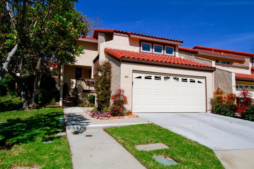 A house in Landmark Yorktown with tan stucco siding and a bright red roof. There is brightly colored floral landscaping and bright green grass with a cement sidewalk entrance.