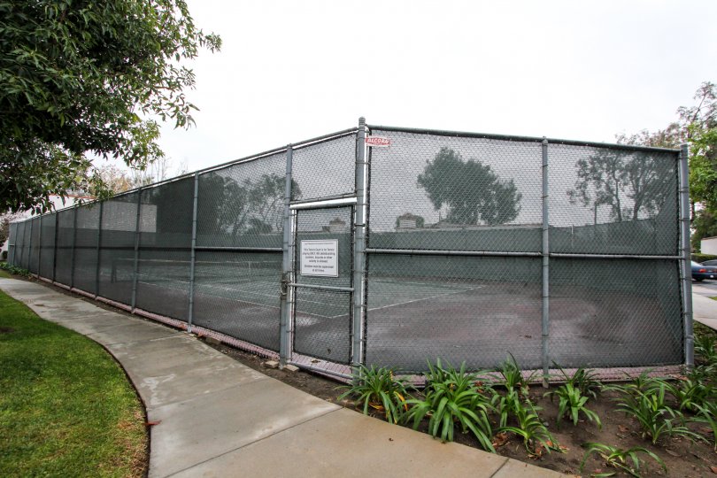 Community tennis court with high meshed wire fence for safety with adjacent sidewalks and green space at Quail Meadows