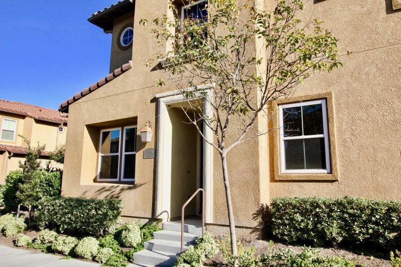 Beige front of house with green shrubs and a young tree, terracotta roof