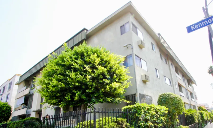 Trees, shrubs, and a black fence surround a white apartment building in the Montclair Kenmore community of Koreatown, California