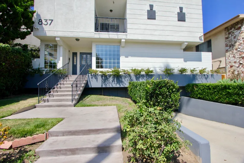 THE BUILDING WITH ENTRANCE STEPS AND A BALCONY ALSO AVAILABLE AT THE MORNING TIME SOME PLANTS NEAR TO THERE.