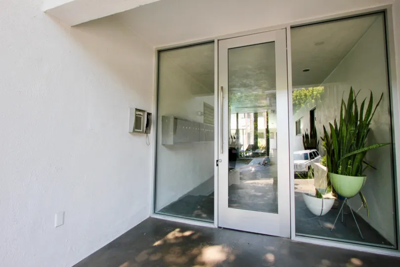 Glass door entranceway with secure intercom and keypad entry. The door leads to a foyer with mailboxes along the wall and potted plants along the opposite wall Reflection suggest the entrance faces a parking area
