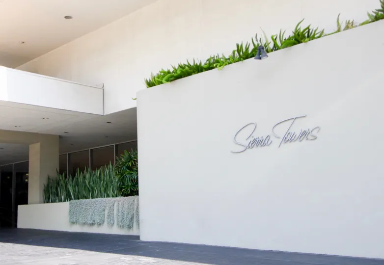 A BOARD THAT REPRESENT A PLACE AND LOOKS LIKE A BUILDING WALL NEAR TO THE PLACE AND SOME PLANTS THERE