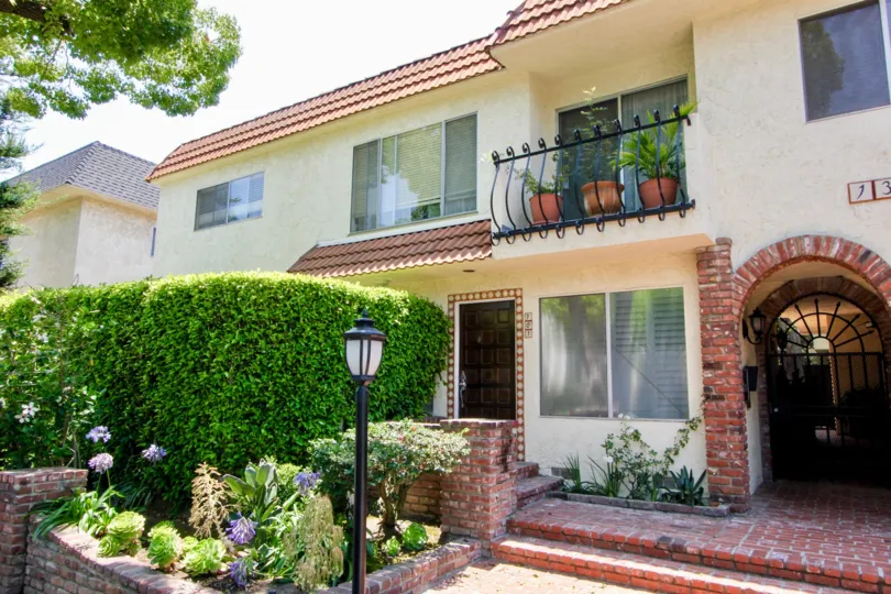 A HOUSE WITH LAMP STAND AND A BALCONY WITH SMALL PLANTS AND ALSO SOME PLANTS IN FRONT OF THE HOUSE
