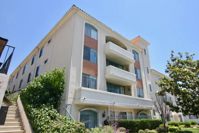 Garden and spanish roof apartment building with steps leading up and lots of windows with front lawn and trees with shrubs
