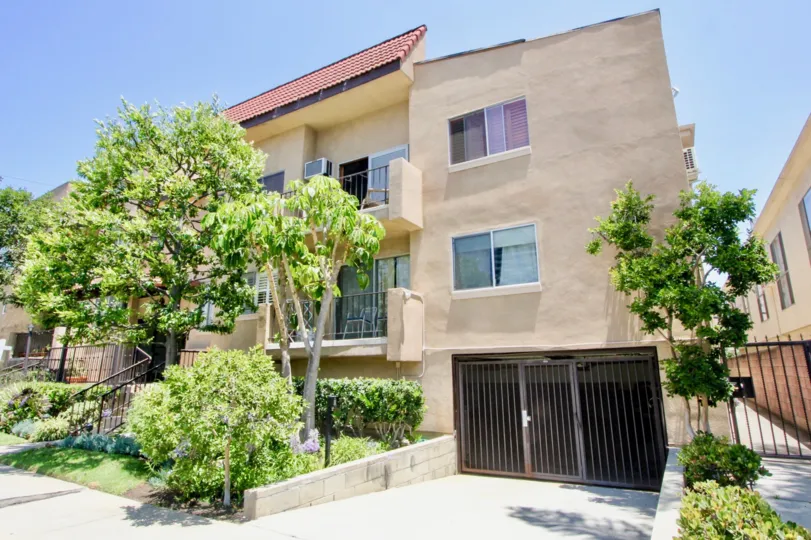 View of apartments with gated entrance to parking; balconies, beautiful green shrubbery landscaping.