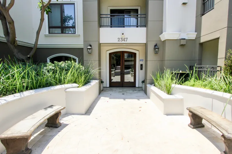 A shady view of Fox Hills Manor, a residential building with an elevated garden and stone benches out front.