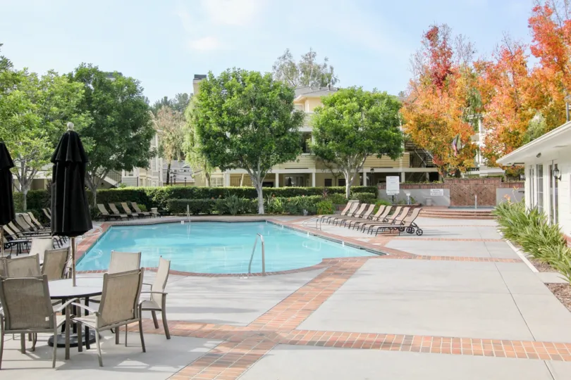 This is a sparkling pool surrounded by beautiful green and orange colored trees with patio tables and umbrellas to open for your comfort and shade