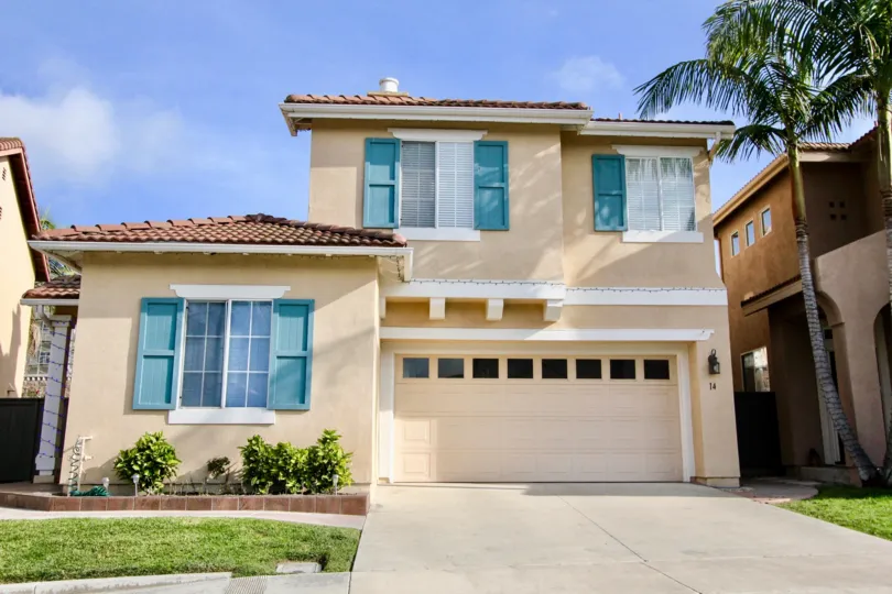 Multi-story stucco home located in the Village Cottages of Aliso Viejo, California. Home includes garage, terra cotta tile roofing, and blue shutters.