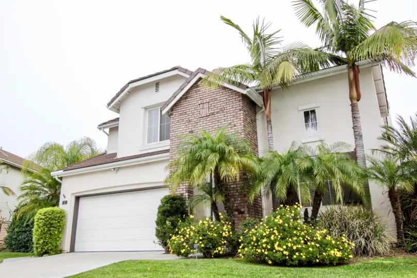 A two-story house with a garage in the Westshore Villas area of Buena Park, CA with palm trees, shrubs, and bushes