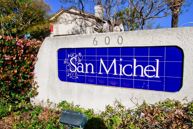 Blue sign to San Michel community with bushes and other plants on a sunny day.