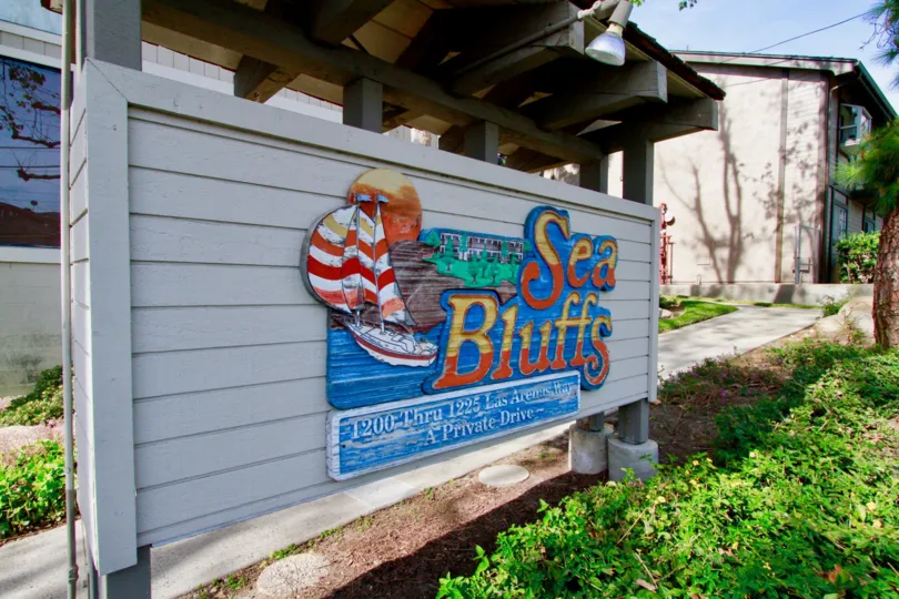 THE CONDOS AT THE SMALL WALL AND IT REPRESENTS SEA BLUFFS WITH BLUE COLOR BACKGROUND BOARD AND A SMALL BUILDING AT THE RIGHT SIDE AND THE SKY LOOKING BLUE AND SOME TREES ALSO AVAILABLE.