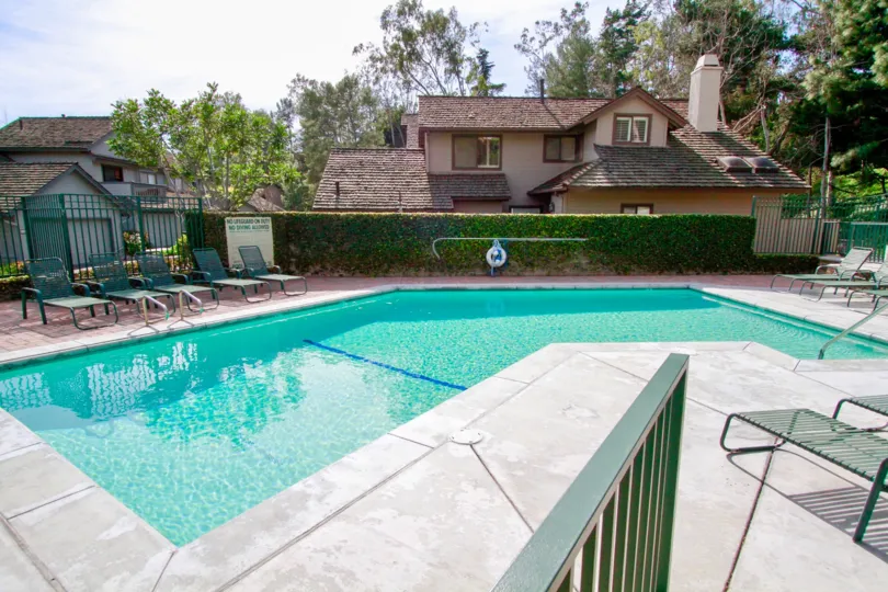 THE SWIMMING POOL WITH SITTING CHAIRS AND THE ENTRANCE GATE LOOKING LIKE GREEN COLOUR AND A SIGN BOARD NEAR TO THE GATE AND SOME TREES ALSO AVAILABLE NEAR TO THE POOL