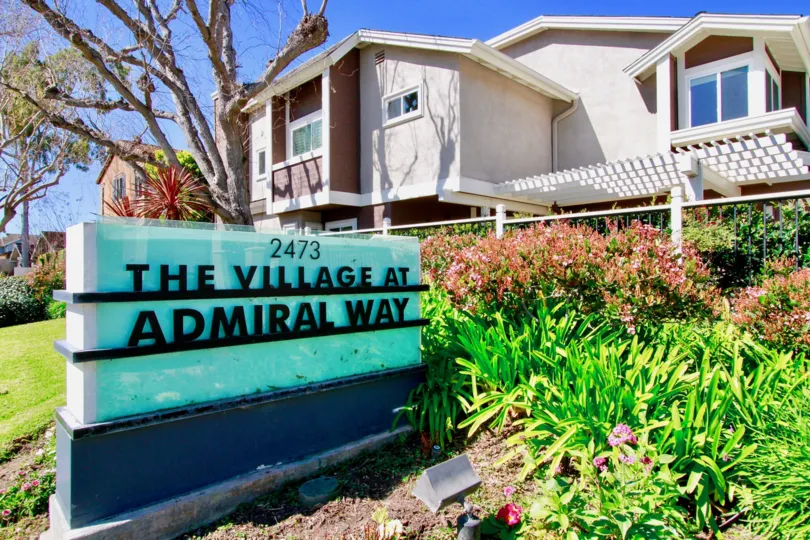 THE BOARD SIGN REPRESENTS THE VILLAGE AT ADMIRAL WAY AND IT LOOK LIKE BLUE BACKGROUND AND THE BUILDING ALSO LOCATED AT THE BACK SIDE OF THE BOARD SOME PLANTS NEAR TO THE SIGN
