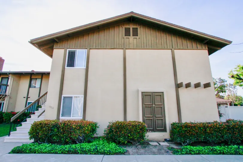 THE BUILDING ALONE WITH A DOOR CLOSED AND TWO GLASS WINDOWS AND ONE LINE WINDOW SOME PLANTS IN FRONT OF THE HOUSE AND STEPS BEHIND THE HOUSE.