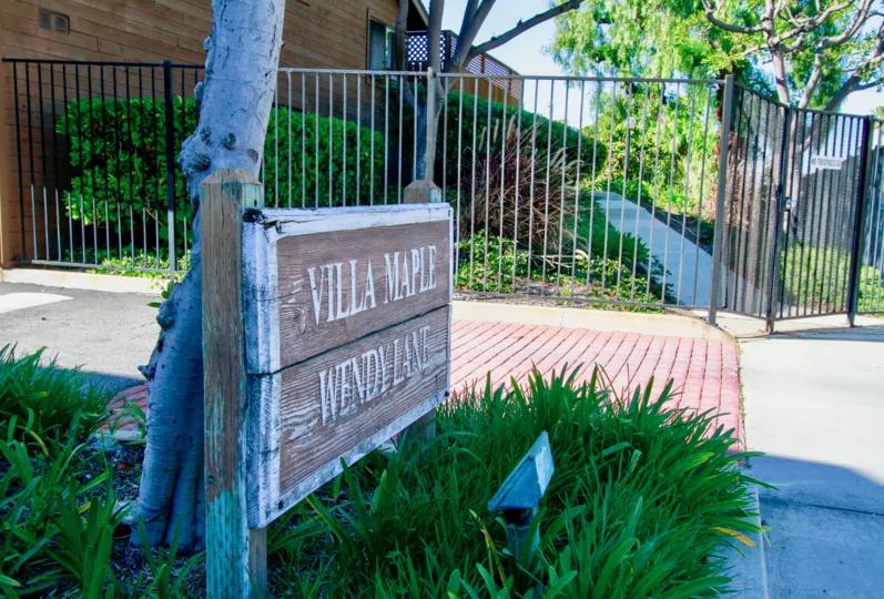 THE BUILDING OF HOUSE WITH THE ENTRANCE GATE AND A SIGN BOARD AT THE FRONT OF THE BUILDING IT LOOKS VILLA MAPLE WENDYLANE AND SOME TREES AND PLANTS SURROUNDING OF THE SIGN