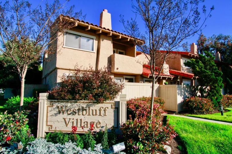 THE PLACE WITH A BOARD SIGN THAT REPRESENTS WESTBLUFF VILLAGE AND SOME GARDENDS WITH FLOWER IN FRONT OF THE HOUSE AND THE HOUSE LOOKING WITH PINK COLOUR AND WHITE COLUR AND THE ROAD WAY ALSO IN FRONT OF THE HOUSE.