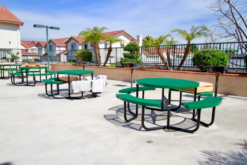 four green round picnic tables with grills placed on concrete slab near houses, towels drying on table benches