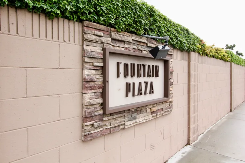 A long tan wall with the sign pictured in tan and brown in the Fountain Plaza community.