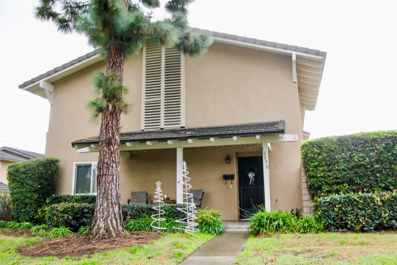Tan housing with decorations outdoors and a tall pine tree in the Green Valley Townhomes community.