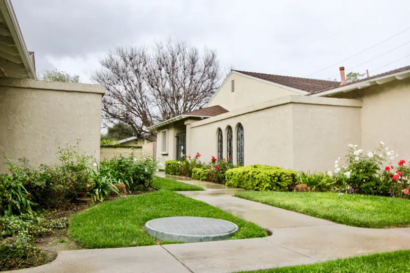 A sidewalk between two homes in the Green Valley Warner Village with grass and plants on each side and a tree in the background.