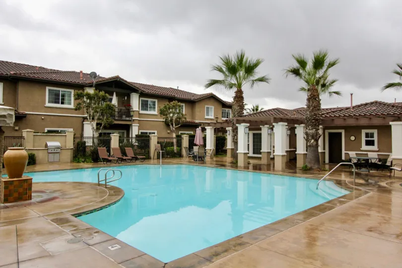 A swimming pool in the Liberty Founder of Village with beautiful buldings, big palm trees, chairs and tables, shrubs.