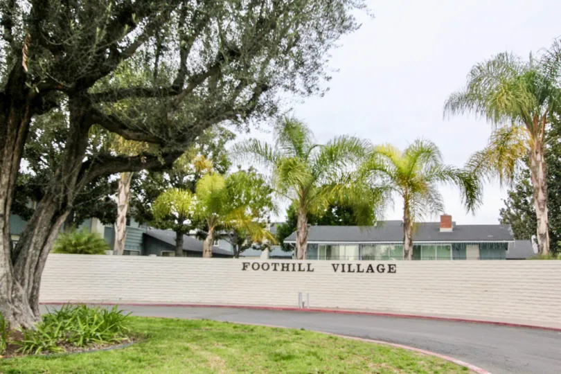 The entrance sign of Foothill Village with palm trees on a cloudy day.