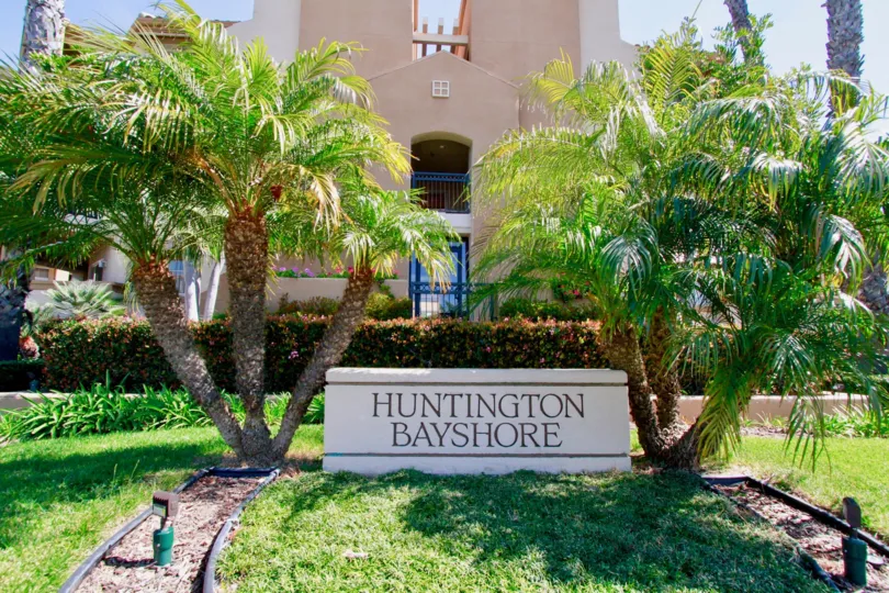 THE HOUSE WITH BLUE COLOUR DOOR AND A BOARD SIGN IN FRONT OF THE HOUSE IT REPRESENTS HUNTINGTON BAYSHORE AND TWO TREES ARE LOCATED AT THE BOARD BOTH SIDES.