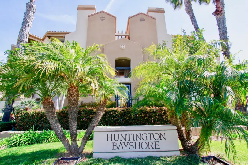 A HOUSE WITH A BOARD OF SIGN IT REPRESENTS HUNTINGTON BAYSHORE AND SOME TREES ALSO BOTH SIDE OF THE BOARD AND A CUTE HOUSE ALSO AT THE BACKSIDE OF THE BOARD SIGN