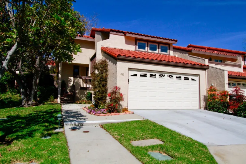 A house in Landmark Yorktown with tan stucco siding and a bright red roof. There is brightly colored floral landscaping and bright green grass with a cement sidewalk entrance.