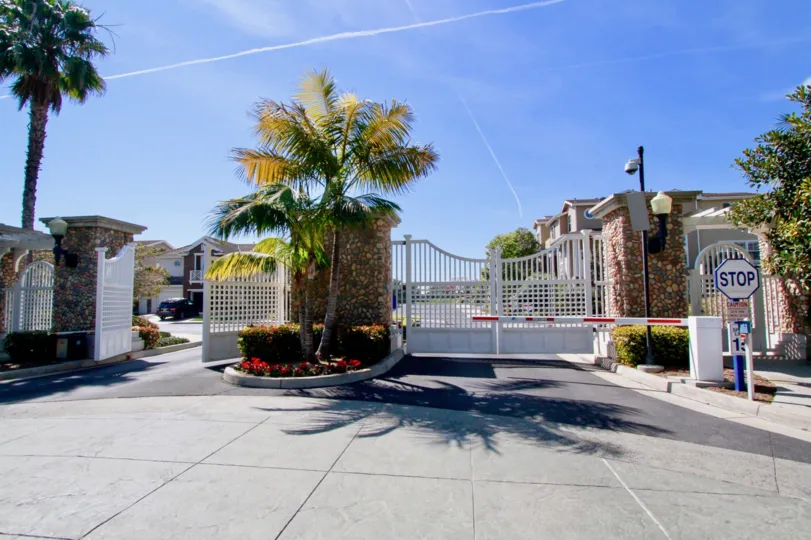 THE MAIN GATE ENTRANCE LOOKING BEAUTIFULLY DECORATE BY THE FLOOR PLANTS AND TREES AND THE SKY ALSLO LOOKING GOOD WITH THE CLOUDS AND A STOP SIGN ALSO AVAILABLE BUT THE APARTMENTS ALSO VIEW AT LITTLE LONG