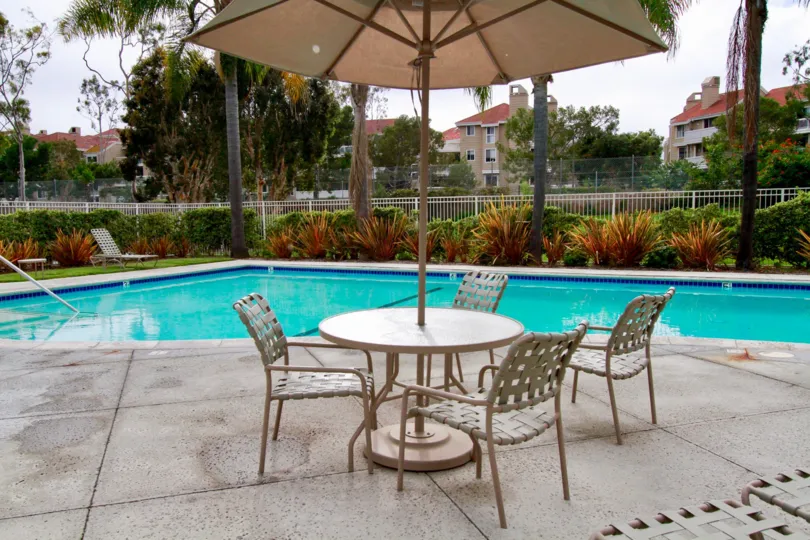 THE SWIMMING POOL WITH UMBRELLA SITTING CHAIRS FOR PROTECT THE SUNSHINE AND SOME PLANTS ALSO SURROUNDING THE POOL.