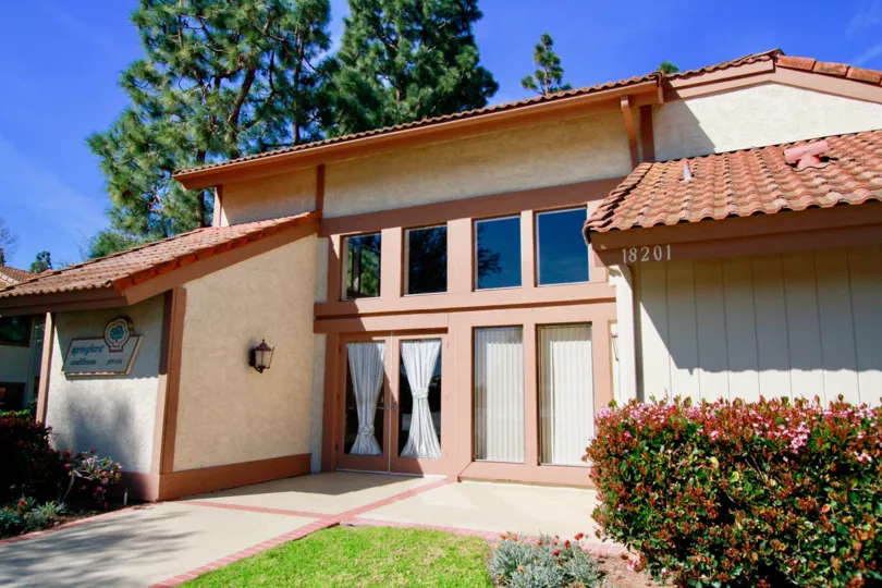 THE SMALL BUNGALOW WITH CUTE DOORS AND SCREES AND WINDOW GLASSES ALL CLOSED AND TOP ROOF SOME PLANTS WITH FLOWERS ALSO NEAR TO THE PLACE.