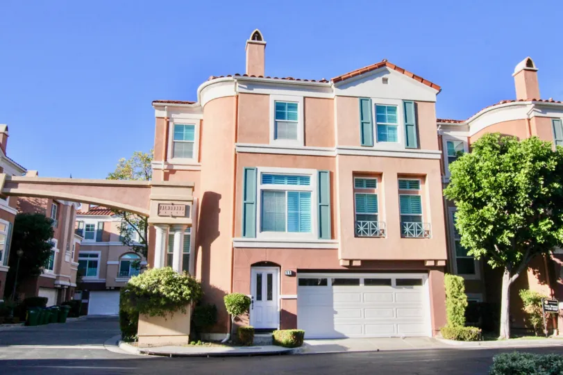 A sunny day in Corte Bella with a large house with pink plaster, big windows and a garage, with a tall tree next to it.