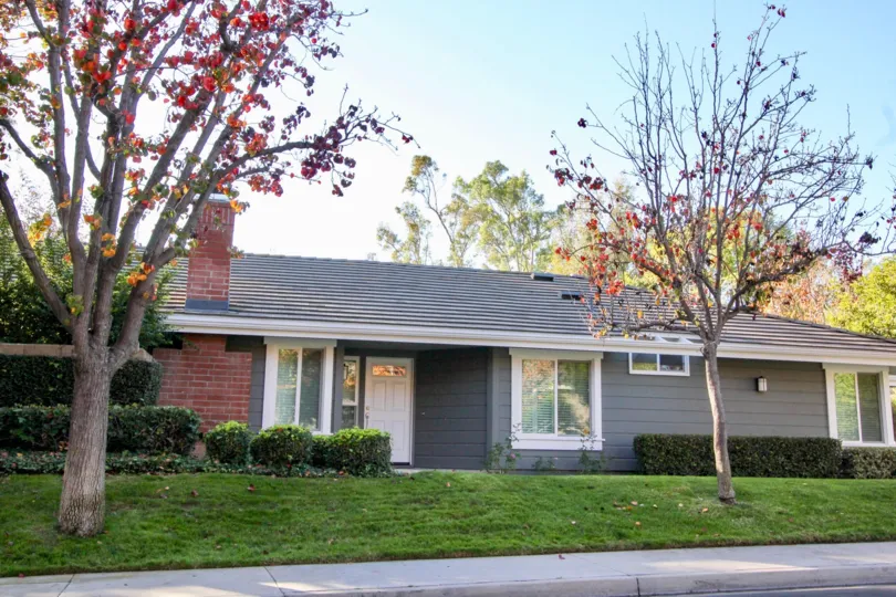 A sunny day in Ridge Garden Homes with a grey bungalow and hedges and trees in front of it.