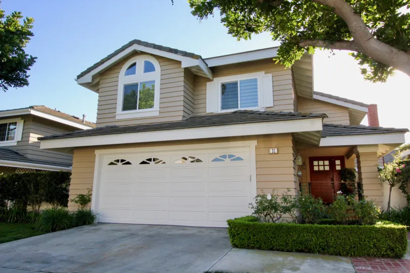 A sunny day in Ridge Garden Homes with a house with a garage for two cars and brown front door and tall tree next to it.