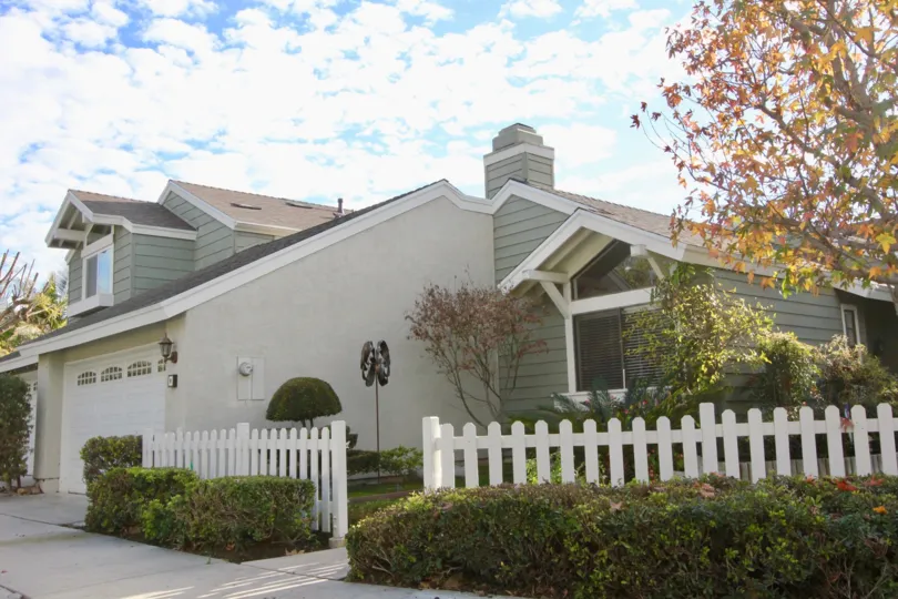 A street view of a home in Windwood Garden Homes with trees, shrubs and white wooden fence