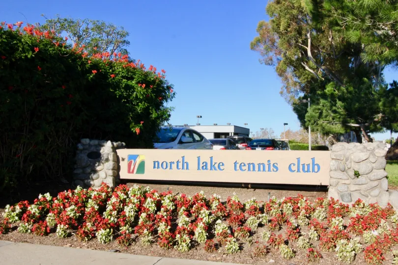 A tennis club signpost in the Woodbridge Grove community surrounded by reddish blooms.