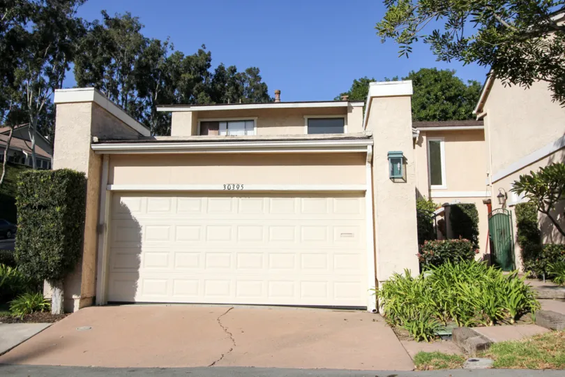 A sunny day by a El Niguel Terrace house with trees behind it and other greenery on each side of the walkway to the front door and the garage door, along with a crack in the driveway concrete