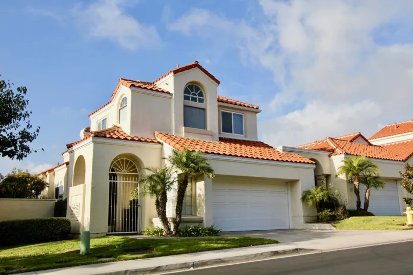 a sunny day in the laguna sur with a house that has the entrance at the left side with a closed parking door in the centre with small trees in front