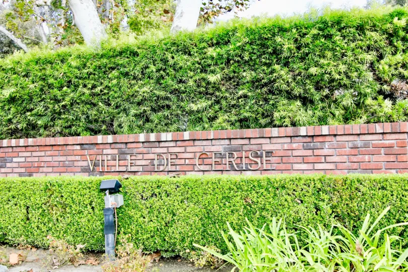 A partly cloudy day by a Ville De Cerise sign on bricks with bushes, trees, and other greenery around