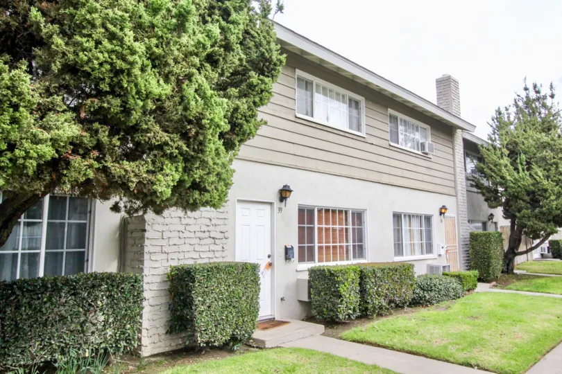 A grey home in The California with bushes and trees on a sunny day