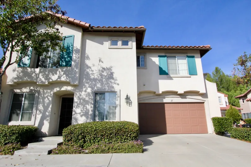 A sunny day by a Fiesta-area house with a tree on one side and bushes on each side of the walkway to the front door and by the garage door