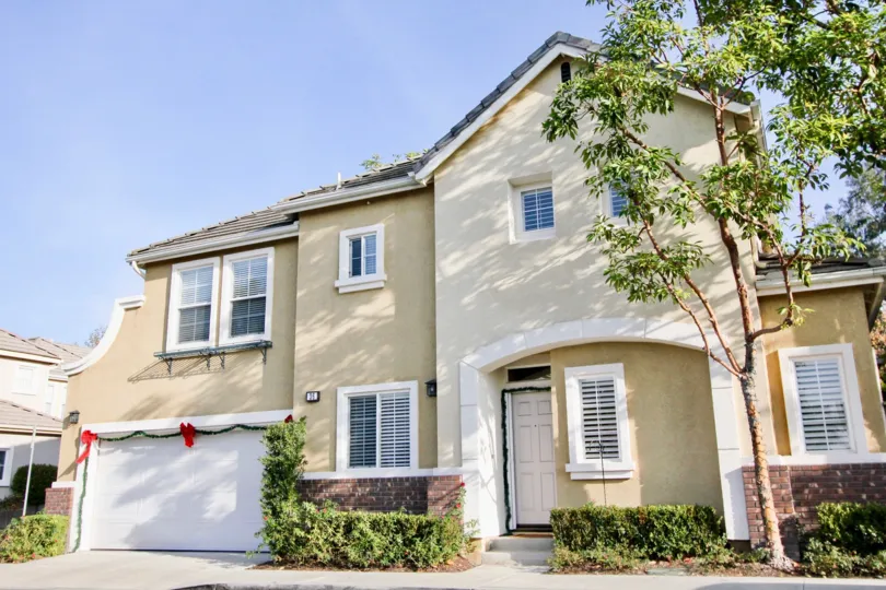 A sunny day by a Magnolia house with Christmas decorations around the garage door, a tree on one side of the house, and a few bushes right in front of it