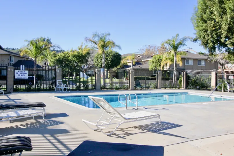 The sparkling blue pool at Casa De Capistrano with pool chairs, tall fencing and palm trees