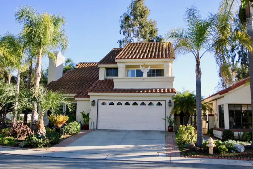 A sunny day at a house at Casitas Del Rio with trees, potted plants, and other plants in front of it, on each side of the driveway