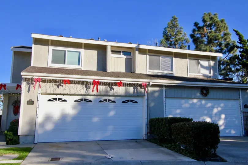 A sunny day by a Mariners Village duplex with Christmas decorations over the garage doors and bushes in between each garage door and trees behind the house