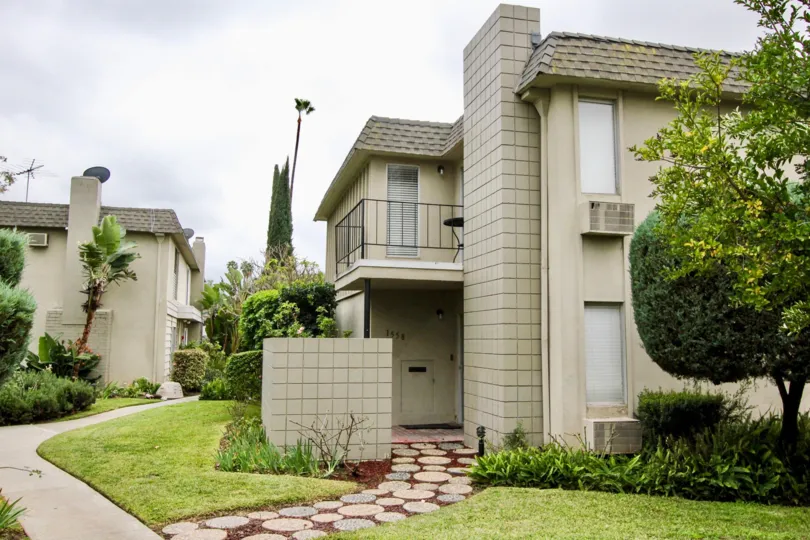 A peaceful suburban home surrounded by several trees and gardens. The house is beige colored and has a balcony.