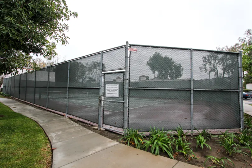 Community tennis court with high meshed wire fence for safety with adjacent sidewalks and green space at Quail Meadows